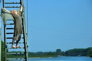 Una donna su una scala apparentemente nel vuoto ha le braccia e il volto protesi verso l'alto e sullo sfondo uno specchi d'acqua circondato da alberi