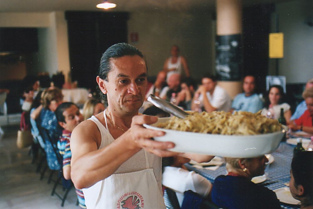 un uomo in canottiera e grembiule che porta un vassoio pieno di spaghetti